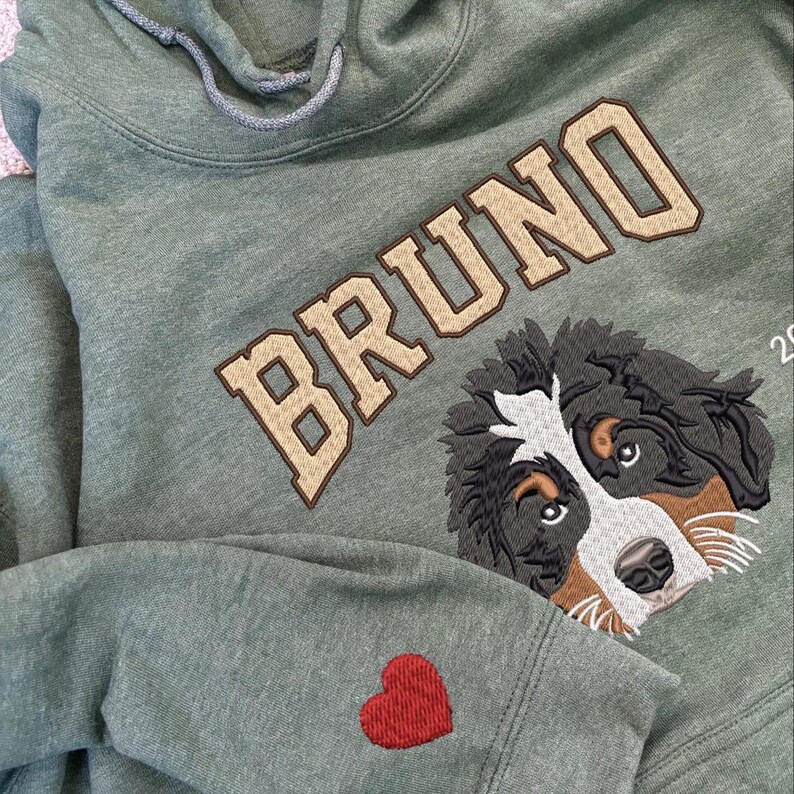 Puede incluir: Una sudadera con capucha verde con el nombre "BRUNO" bordado en beige en la parte delantera. Un perro de monta&ntilde;a bern&eacute;s est&aacute; bordado debajo del nombre. Un coraz&oacute;n rojo est&aacute; bordado en la manga izquierda.