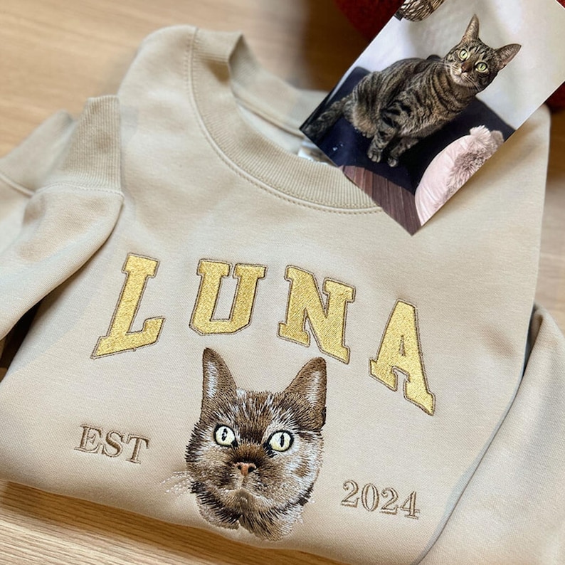 Puede incluir: Una sudadera beige con bordado dorado que dice "LUNA EST 2024". El bordado tambi&eacute;n incluye la cara de un gato.