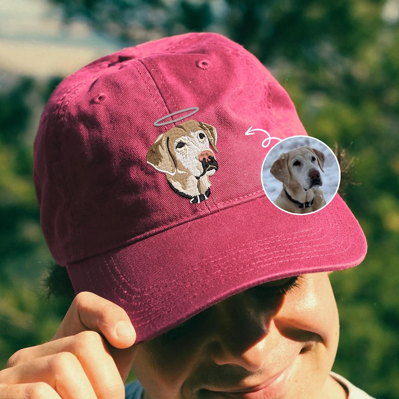 May include: A burgundy baseball cap with an embroidered image of a dog with a halo. The dog is a light brown colour with a white chest and a black nose.
