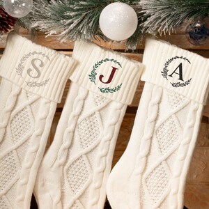 May include: Three cream-colored Christmas stockings with cable knit design. Each stocking has a different initial: "S", "J", and "A", inside a wreath. The stockings are hanging near Christmas decorations, including ornaments and pine branches.