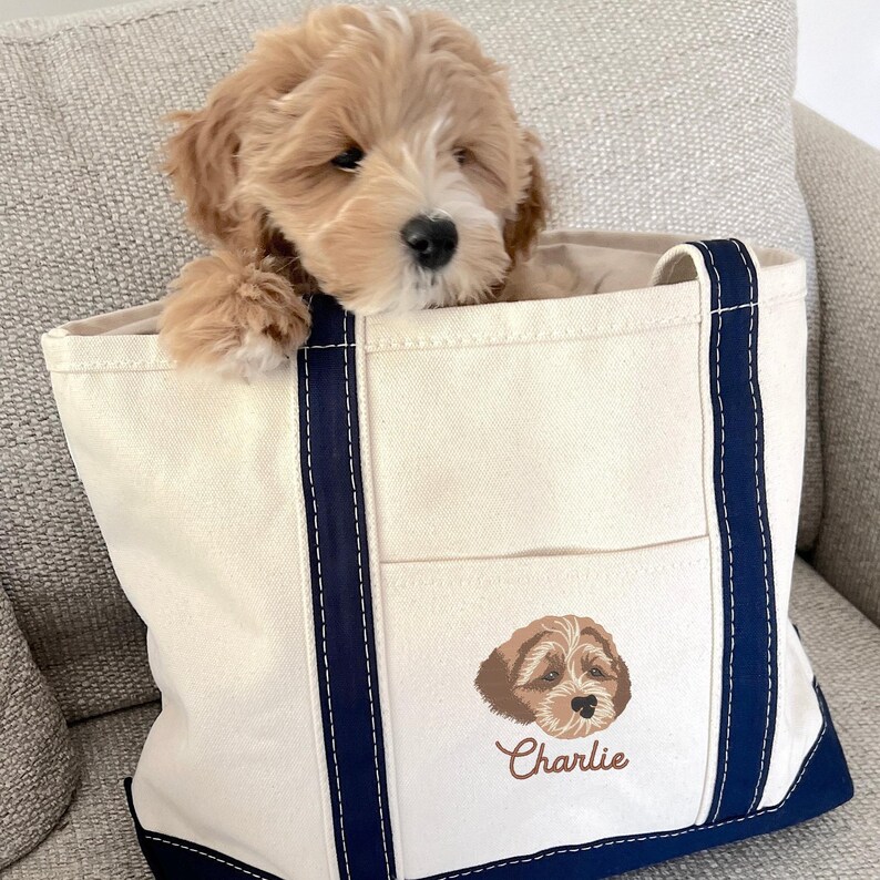 May include: A cream-colored canvas tote bag with navy blue trim and a large pocket on the front. The pocket features an embroidered image of a brown and white dog with the name "Charlie" embroidered below.