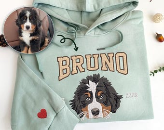 May include: A light green hoodie with a brown and white dog's face embroidered on the front. The dog's name, "BRUNO", is embroidered in brown letters above the dog's face. The year "2023" is embroidered in small letters on the right side of the dog's face. A small red heart is embroidered on the left sleeve of the hoodie.