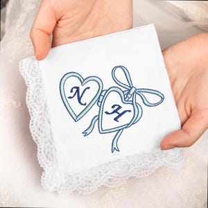 May include: White handkerchief with a delicate lace trim, featuring two embroidered blue hearts. Each heart contains a letter, with ribbons and a bow adding detail. A personalized wedding or anniversary gift.