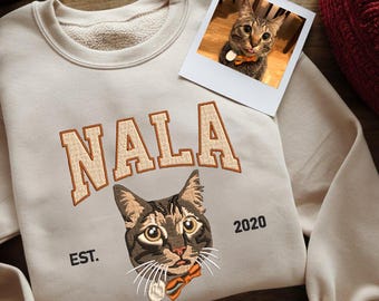 Könnte beinhalten: Ein beiger Pullover mit einem braunen Katzen-Stickbild auf der Vorderseite. Die Katze trägt eine Fliege und der Pullover sagt "NALA EST. 2020".