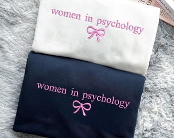 May include: Two sweatshirts with the text "women in psychology" and a pink bow embroidered on each. The top sweatshirt is white and the bottom sweatshirt is black.