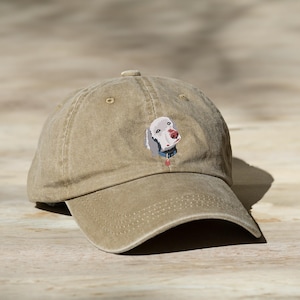May include: A tan cotton baseball cap featuring an embroidered image of a Weimaraner dog. The dog is looking at the camera with its head tilted slightly to the side.