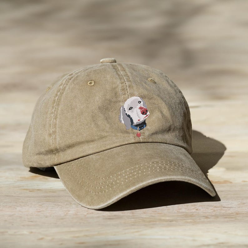 May include: A tan cotton baseball cap featuring an embroidered image of a Weimaraner dog. The dog is looking at the camera with its head tilted slightly to the side.