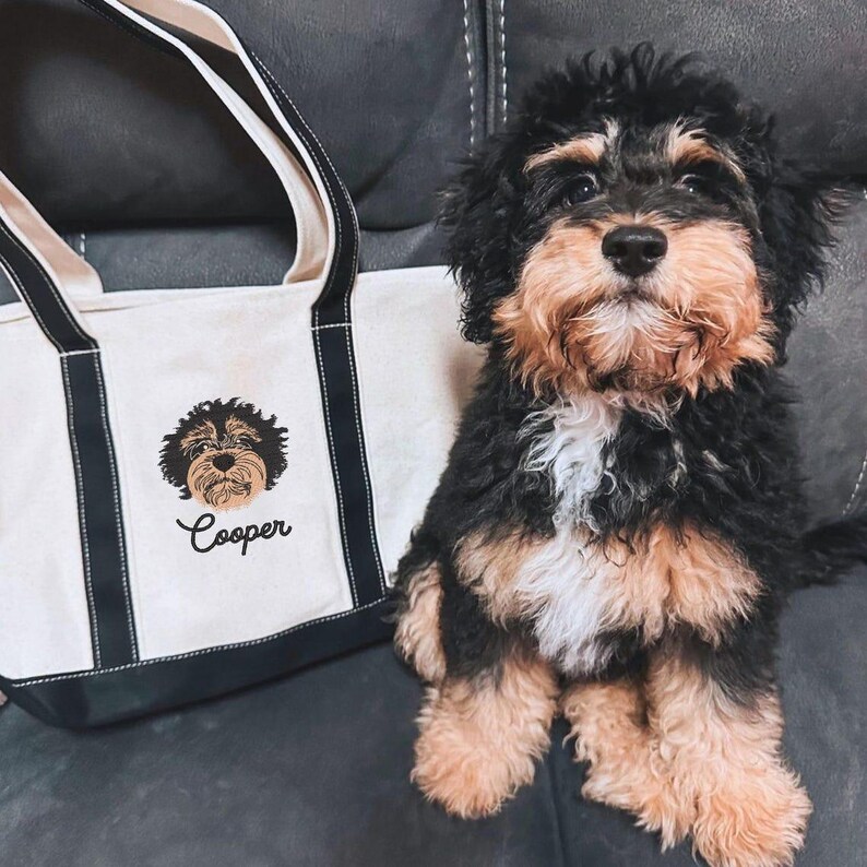 May include: A white canvas tote bag with black trim and a black embroidered image of a dog with the name "Cooper" underneath. A black and brown dog is sitting on a gray couch.