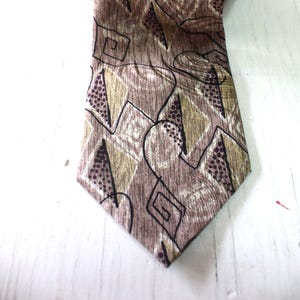 May include: A brown tie with an abstract geometric pattern. The design includes shapes in shades of brown, tan, and purple, outlined in black. The tie is made of a textured fabric.