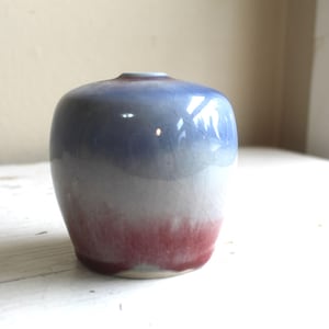 以下が含まれることがあります： グラデーションカラーの小さな丸みを帯びたセラミック花瓶。上部は濃い青色で、中央は白に変わり、赤紫色のベースで終わります。花瓶は滑らかで光沢のある仕上がりで、上部に小さな開口部があります。