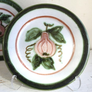 May include: White ceramic plate with a green rim and a hand-painted design. The design features a pink fruit with green leaves and brown vines. A thin red-brown ring encircles the fruit. The plate is displayed on a clear stand.