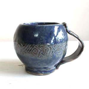May include: A handmade, blue ceramic mug with a rounded body and a curved handle. The mug features a textured, geometric pattern around the middle. The glaze has a glossy finish, and the mug is suitable for hot or cold beverages.