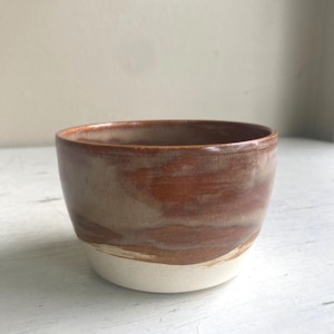 Op de afbeelding: Een kleine keramische kom met een ronde vorm, voorzien van een glad, glanzend bruin glazuur aan de bovenzijde dat overgaat in een matte, gebroken witte basis. De binnenzijde van de kom heeft een egale bruine afwerking met subtiele kleurnuances.