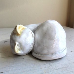 May include: A white ceramic sculpture of a sleeping cat with a yellow ear. The cat is curled up in a ball with its head resting on its paws.
