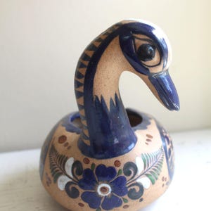 Może przedstawiać: Ceramiczna figurka łabędzia z niebieską głową i szyją oraz beżowym korpusem zdobionym niebieskimi wzorami kwiatowymi. Szyja łabędzia jest zakrzywiona, a korpus okrągły. To przedmiot dekoracyjny.