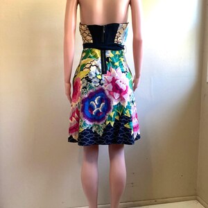 May include: A strapless mini dress featuring a vibrant floral pattern with large pink, purple, and blue flowers on a multi-colored background. The bodice includes a solid black panel at the center back with a visible vertical zipper closure.