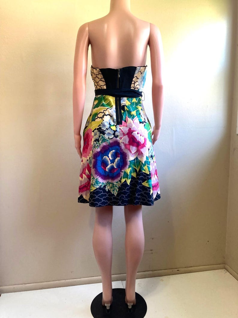 May include: A strapless mini dress featuring a vibrant floral pattern with large pink, purple, and blue flowers on a multi-colored background. The bodice includes a solid black panel at the center back with a visible vertical zipper closure.