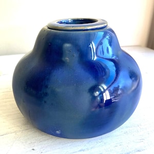 Può includere: Un vaso di ceramica blu con corpo arrotondato e apertura stretta. Il vaso ha una finitura lucida e una leggera forma a pera. Il colore è un blu intenso e ricco con sottili variazioni di tonalità. Il bordo ha un accento dorato.