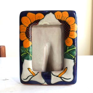 May include: Rectangular ceramic picture frame with a blue border. The frame is decorated with orange sunflowers and white calla lilies. The frame has a neutral-colored interior for displaying a photograph.