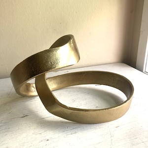 May include: A gold-coloured sculpture featuring a continuous, twisted ribbon-like band that forms an open, circular structure. The surface has a metallic finish with subtle texture, creating a fluid, looping shape that rises and curves.
