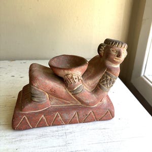 Puede incluir: Una escultura de arcilla color terracota que representa una figura reclinada sobre una base rectangular. La figura sostiene un pequeño cuenco circular frente al torso. La base presenta un patrón triangular tallado y la figura tiene detalles grabados en los brazos y la cabeza.