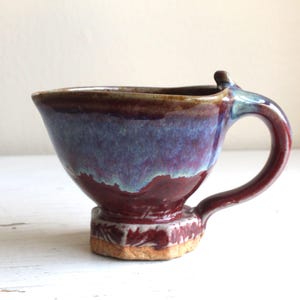 Könnte beinhalten: Eine kleine, handgefertigte Keramiktasse mit einem einzigartigen, asymmetrischen Henkel. Die Tasse ist in Violett-, Blau- und Brauntönen glasiert und hat einen strukturierten Boden.