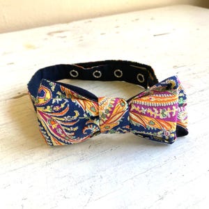 Op de afbeelding: Een marineblauwe vlinderdas met een paisley patroon in tinten oranje, roze en geel. De vlinderdas heeft een zwarte verstelbare nekband met metalen drukknopen, geschikt voor verschillende halsmaten. De vlinderdas is een modeaccessoire.