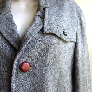 May include: A grey tweed jacket with a brown leather collar and brown leather buttons. The jacket has a flap pocket on the left side.