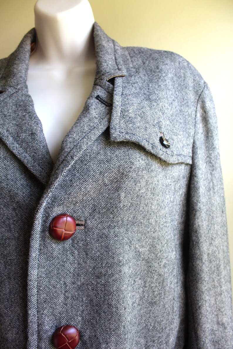 May include: A grey tweed jacket with a brown leather collar and brown leather buttons. The jacket has a flap pocket on the left side.