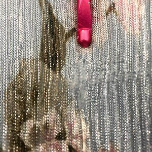Op de afbeelding: Close-up van een roze make-up kwast op een zilver-gouden, getextureerde stof. De kwast is naar beneden gericht, de haren zijn buiten beeld. De stof heeft een glanzende, metallic uitstraling.
