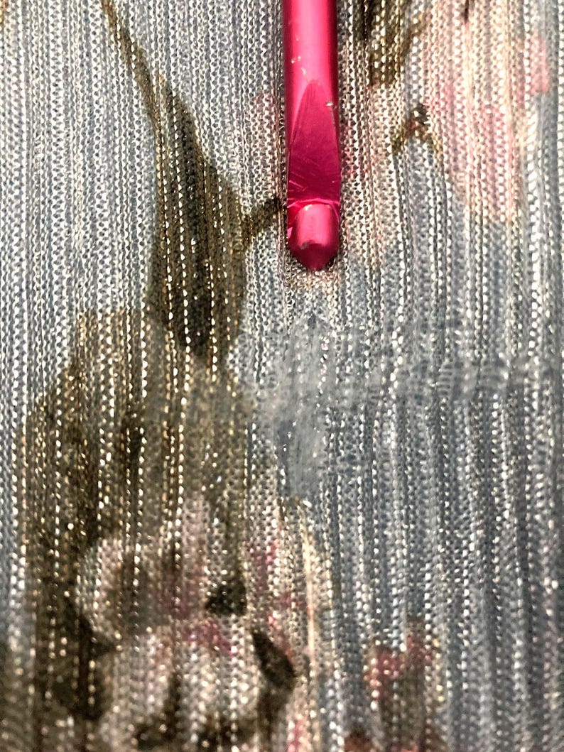 Op de afbeelding: Close-up van een roze make-up kwast op een zilver-gouden, getextureerde stof. De kwast is naar beneden gericht, de haren zijn buiten beeld. De stof heeft een glanzende, metallic uitstraling.