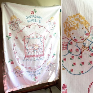 May include: White cotton baby blanket with embroidered angels and the text "Guardian Angels Will Watch Over Me". The angels are wearing pink and blue dresses with floral accents.