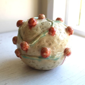 May include: A spherical ceramic ornament with a textured, cream-coloured surface. It has evenly spaced, reddish-orange, rounded knobs. A pale blue, wavy line encircles the sphere. Likely a decorative item or art piece.