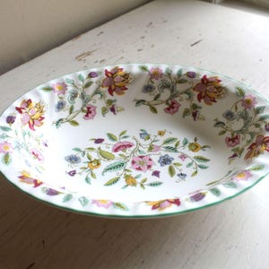 May include: Oval ceramic serving dish with a white base and a floral pattern. The design features pink, yellow, and purple flowers with green leaves and stems. The rim is scalloped and outlined with a green border.