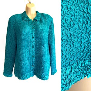 Könnte beinhalten: Türkisfarbenes Button-Down-Shirt mit strukturiertem, geknittertem Stoff. Das langärmlige Oberteil hat einen Kragen und kleine, passende Knöpfe auf der Vorderseite. Eine Nahaufnahme zeigt die einzigartige, wellenartige Textur des Materials.