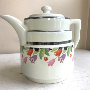 May include: Cream-colored ceramic teapot with a floral design. The teapot features a lid, spout, and handle. The base is decorated with a band of colorful flowers, including orange, purple, and pink tulips. Silver trim accents the body.