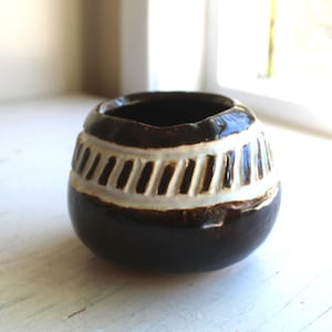 May include: A small, round ceramic vase with a dark brown base and a band of white, vertical stripes around the middle. The interior of the vase is dark, and the rim is slightly uneven. The vase is sitting on a white surface.
