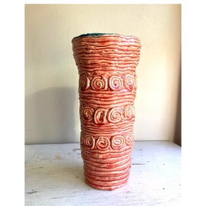 Puede incluir: Un jarrón cilíndrico de cerámica, alto, con un exterior texturizado de color rojo-naranja. El jarrón presenta crestas horizontales y diseños en espiral. El borde tiene un toque de turquesa. El jarrón se exhibe sobre una superficie blanca.