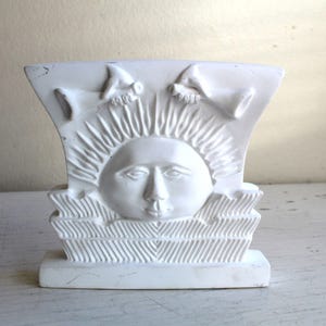 Könnte beinhalten: Weiße Keramikskulptur eines Sonnen Gesichts mit stilisierten Strahlen, flankiert von zwei stilisierten Vögeln mit ausgebreiteten Flügeln. Die Skulptur hat eine strukturierte Oberfläche und eine Basis.