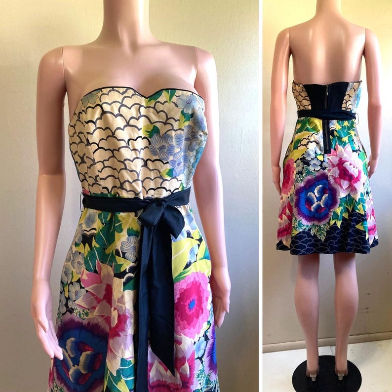 May include: A strapless knee-length dress featuring a vibrant floral pattern with large pink, blue, and yellow blossoms against a cream and black background. The garment includes a solid black fabric waist tie and a visible black zipper on the back.