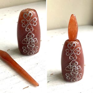 May include: A reddish-brown glass bottle with a silver floral design. The bottle has a long, oval shape and a matching stopper. The stopper is a similar color and shape. The bottle is likely used for perfume or essential oils.