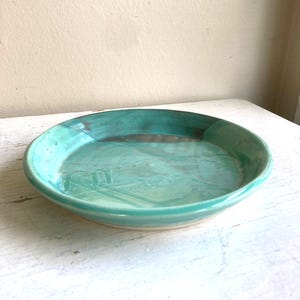 Puede incluir: Un plato de cerámica hecho a mano con un esmalte turquesa. El plato tiene una forma redonda y poco profunda con un borde ligeramente elevado. El interior tiene un aspecto moteado, con variaciones en el color turquesa. El plato es adecuado para servir alimentos.