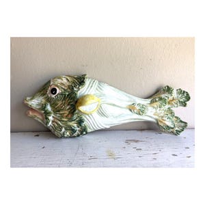 Può includere: Una scultura da parete in ceramica a forma di pesce, realizzata a partire da un cespo di sedano. L'oggetto presenta foglie testurizzate verdi e bianche che formano il corpo e la coda, con una fetta di limone gialla che funge da occhio.