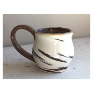 May include: Cream-colored ceramic mug with a brown handle and dark brown brushstroke design. The interior rim is also brown. The mug has a rounded shape and sits on a white surface. A handmade pottery item.