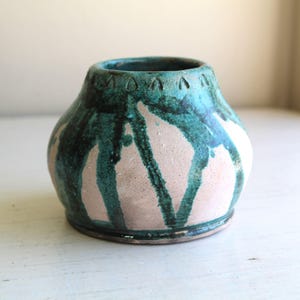 Puede incluir: Un pequeño jarrón de cerámica hecho a mano con un cuerpo redondeado y una abertura ancha. El jarrón presenta un esmalte turquesa y aguamarina con acentos goteados sobre una base rosa claro. El borde tiene un patrón decorativo.