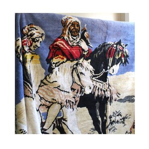 Puede incluir: Un tapiz que representa a un hombre montando un caballo, con una mujer de pie detrás de él. El hombre lleva una túnica roja y blanca, y el caballo es negro con marcas blancas. El tapiz tiene un fondo azul y está tejido con detalles intrincados.