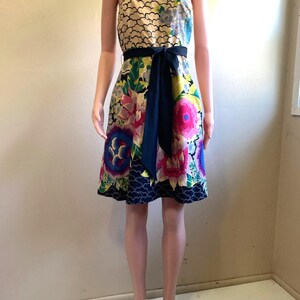 May include: A strapless knee-length dress featuring a cream-colored bodice with a black scalloped pattern, a vibrant floral print skirt in shades of pink, blue, and green, and a solid black fabric sash tied at the waist with a front bow.