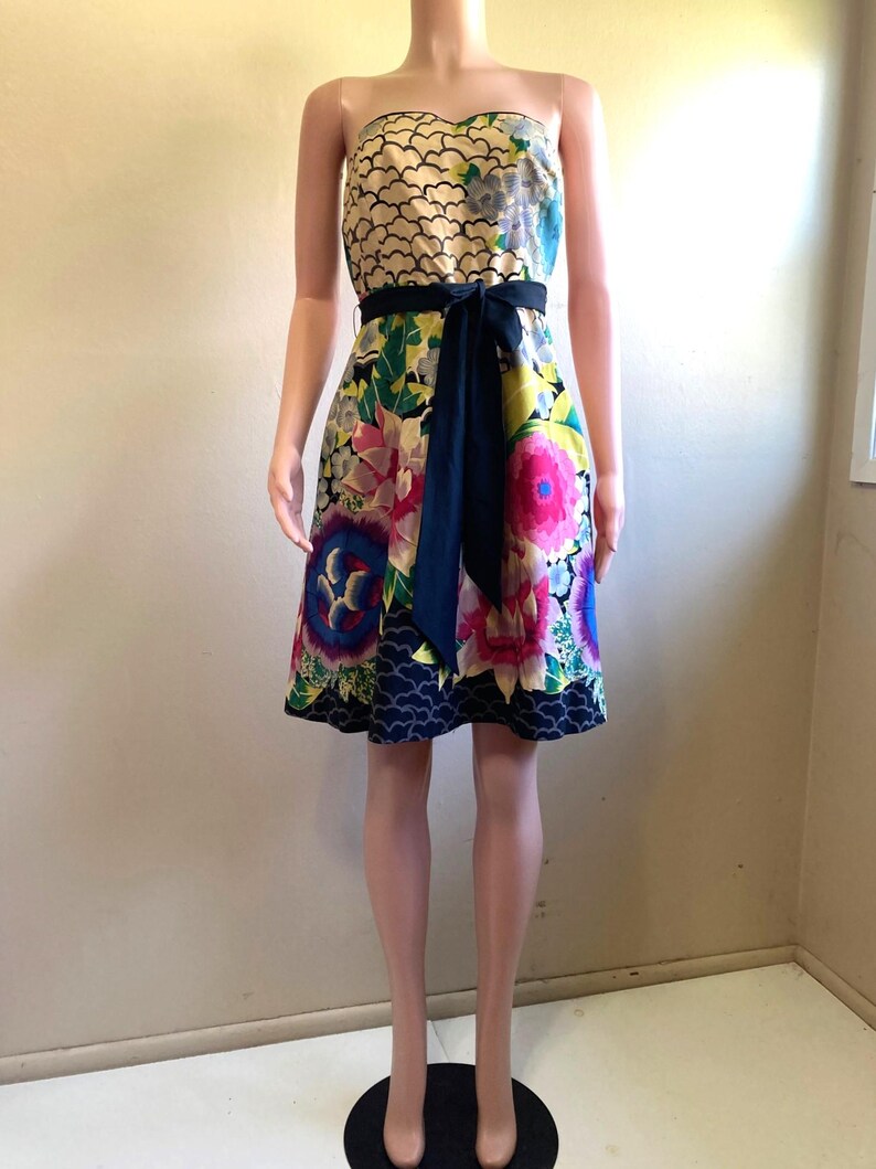 May include: A strapless knee-length dress featuring a cream-colored bodice with a black scalloped pattern, a vibrant floral print skirt in shades of pink, blue, and green, and a solid black fabric sash tied at the waist with a front bow.