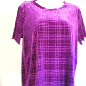 May include: A short-sleeved, purple velvet top with a plaid pattern. The shirt has a round neckline and a slightly scalloped hem and sleeve edges. The fabric has a soft, lustrous appearance.