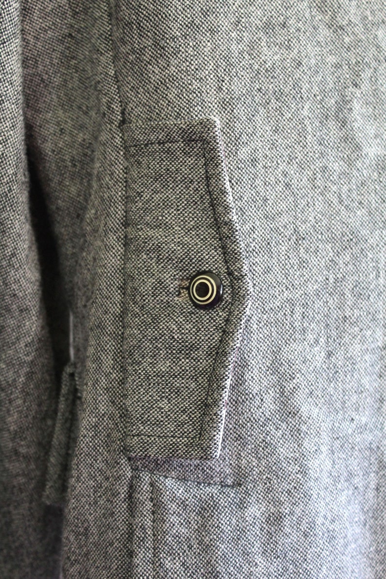 May include: A close-up of a grey tweed jacket with a black button on a flap pocket.